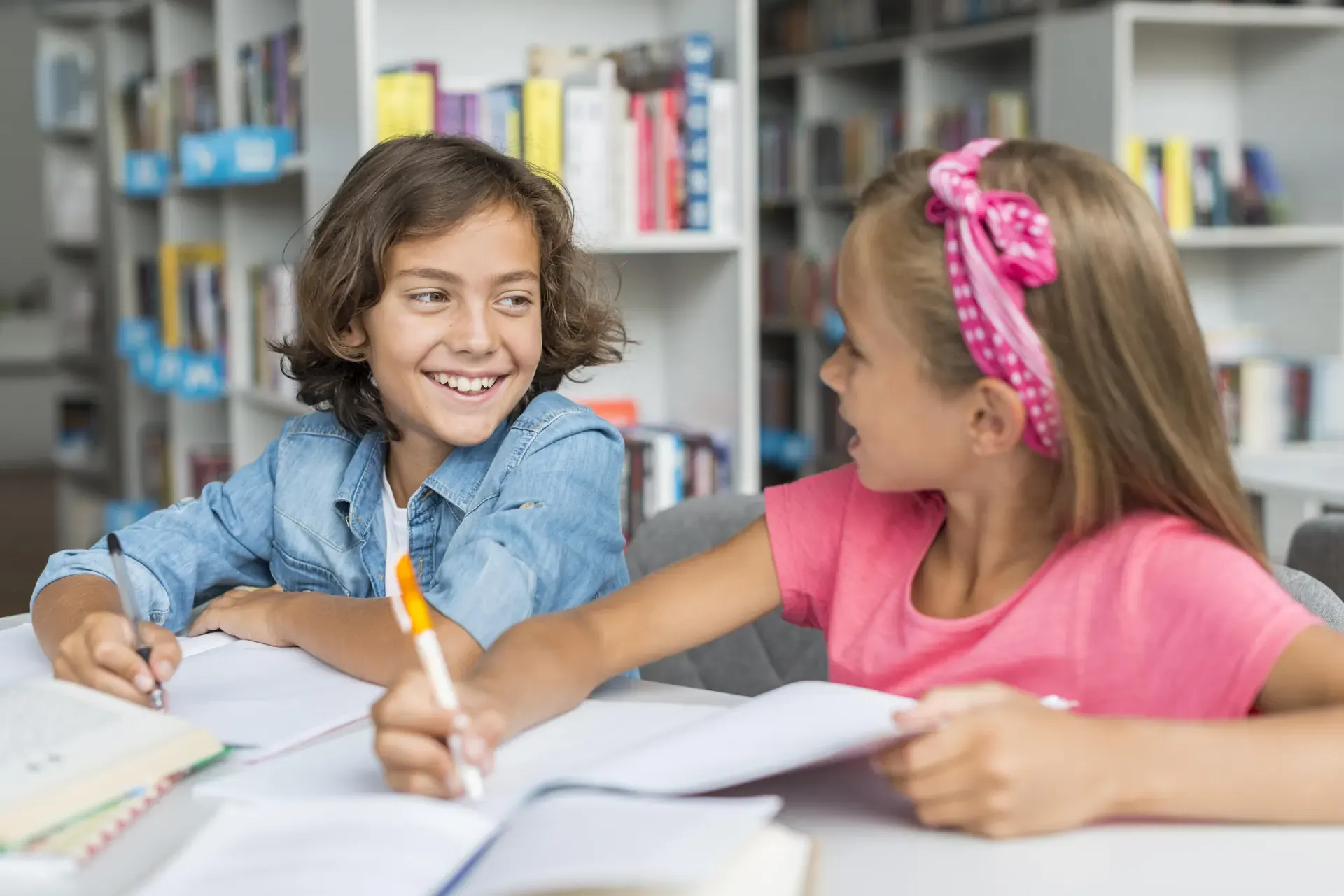 Zwei Kinder sitzen in der Bibliothek und lächeln sich beim Lernen an