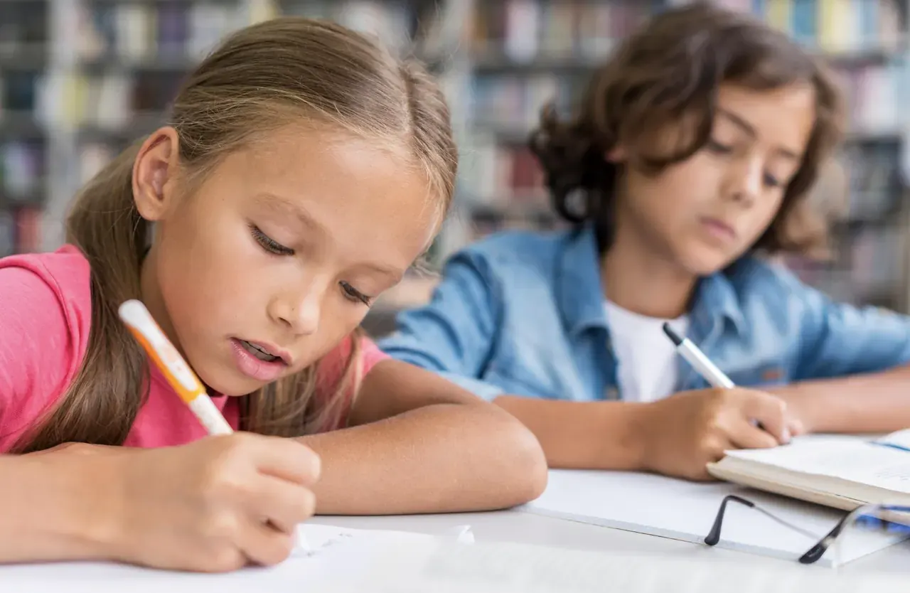 Zwei Kinder schreiben nebeneinander an einem Tisch in der Bibliothek